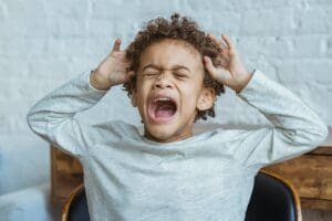 Young boy showing frustration indoors with hands on head, eyes closed, mouth open.