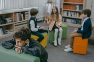 A sad child isolated in a school library while peers chat in the background.