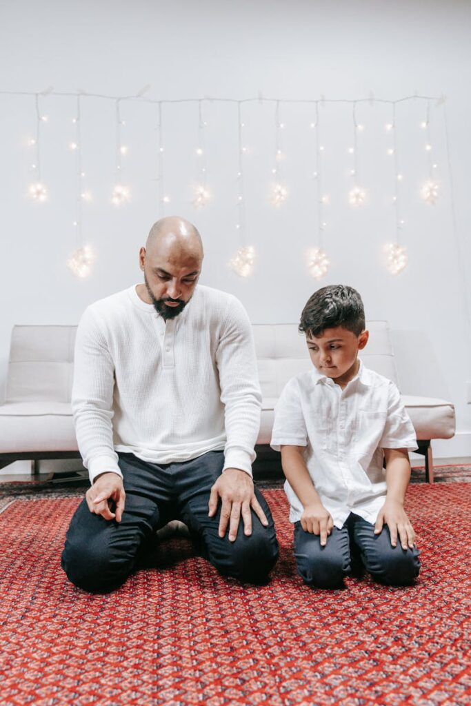 Man and son engaged in prayer on a red carpet with decorative lights indoors.