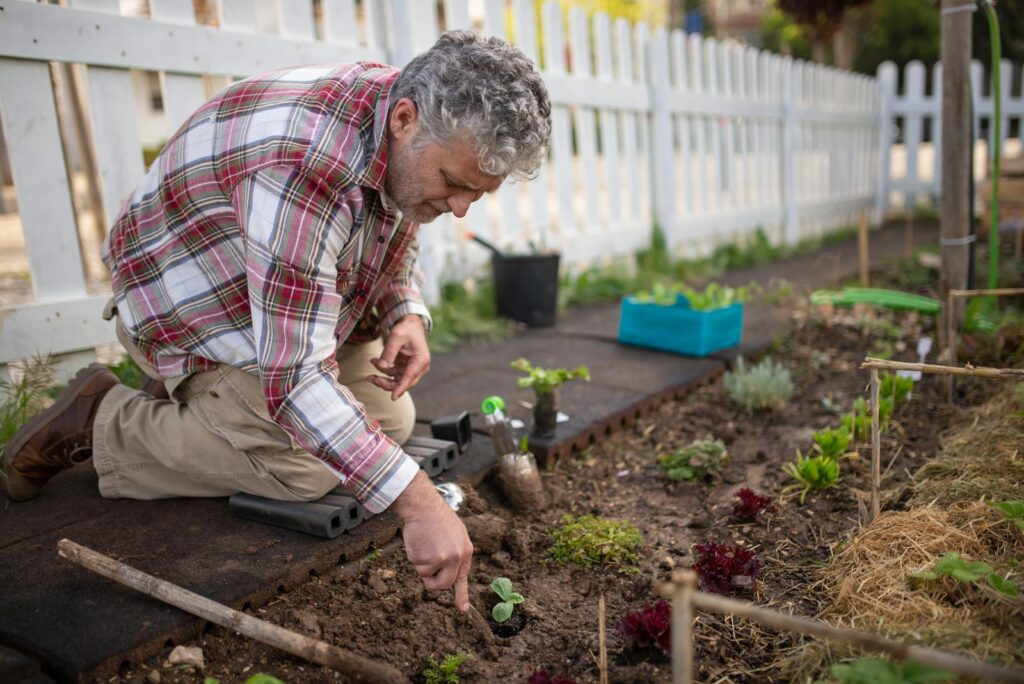 An elderly man kneels to plant in a vibrant garden, surrounded by tools and greenery - AboutBoulder.com