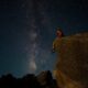 person sitting on edge of large rock under starry night sky